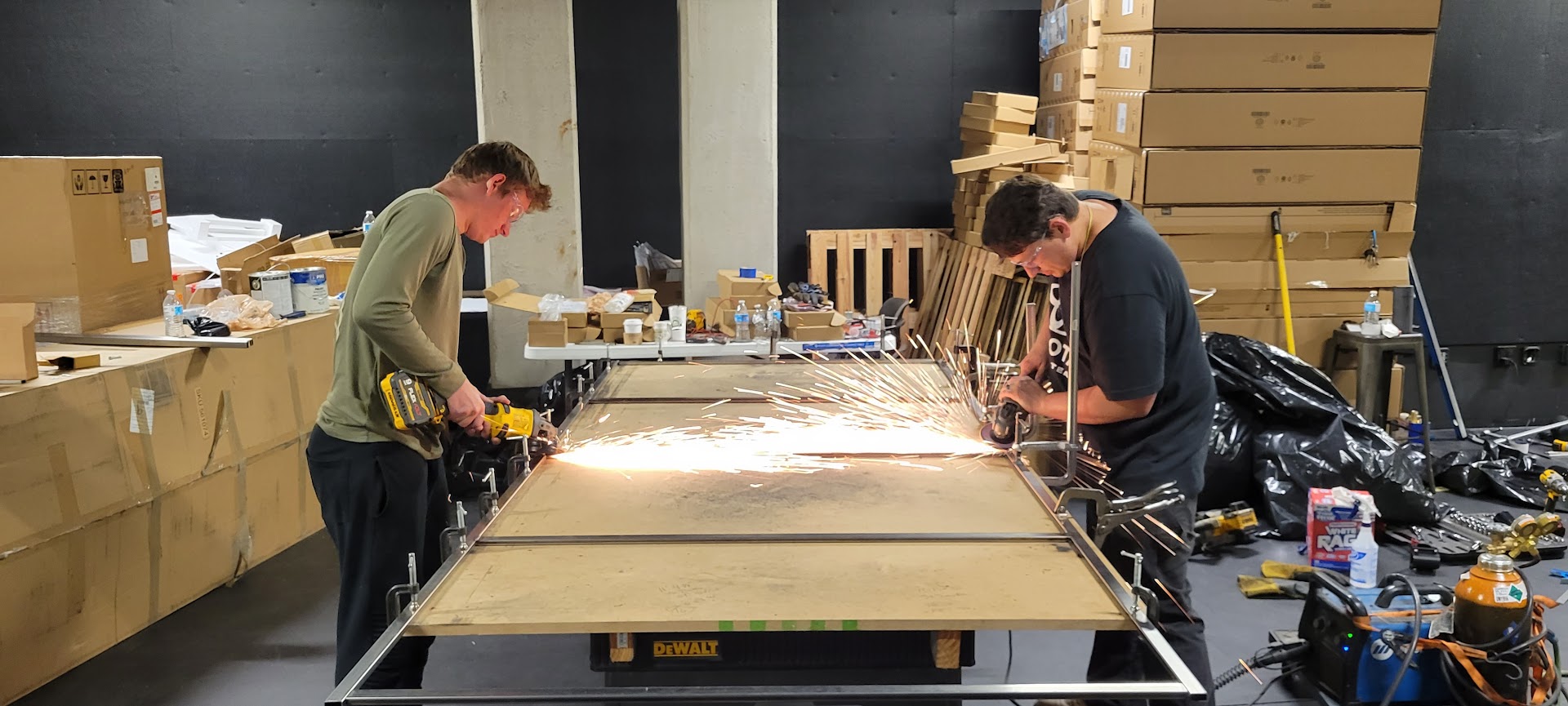 Two crew members grinding a fabricated steel frame as sparks meet across the worktable
